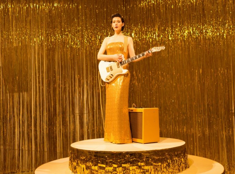 A white woman with dark hair standing on a gold platform, surrounded by gold tinsel, wearing a gold dress, and holding a guitar.