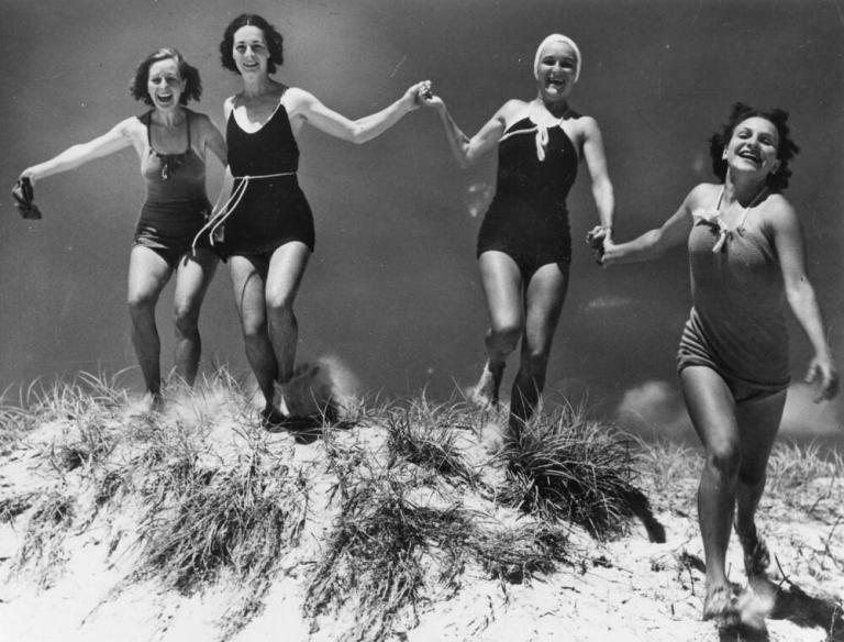 Four friends in 1930s bathing costumes running over a sand dune