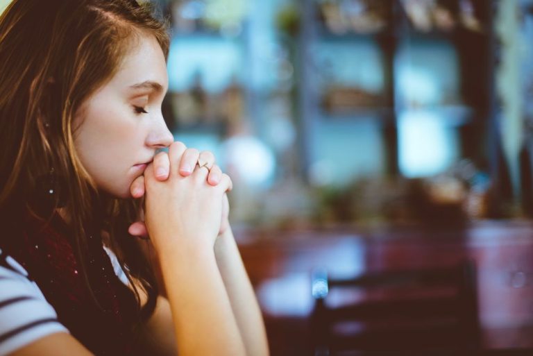 A white woman sits with her head bowed and eyes shut, praying