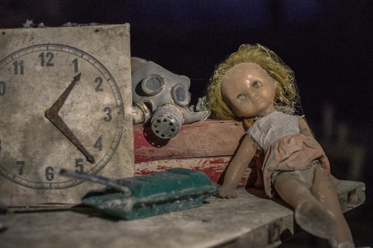 A broken doll sits next to a gas mask and pile of books.