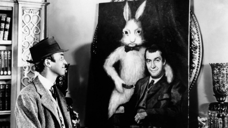 Black and white photo of a man staring at a painting of himself sitting next to a giant rabbit with a bowtie.
