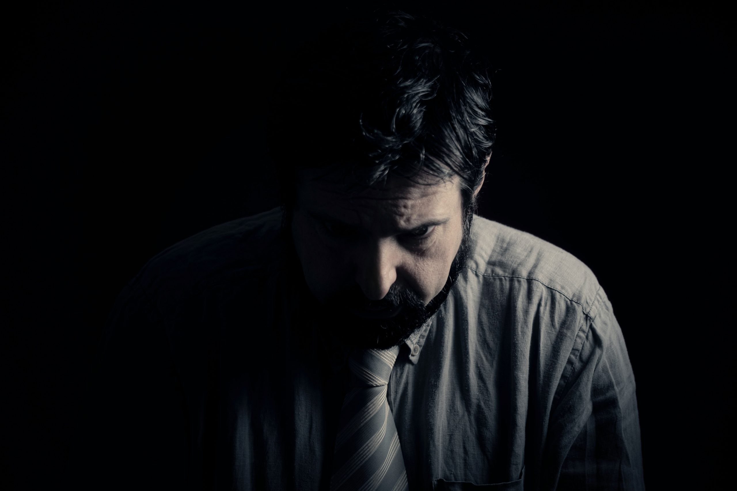 A man in gray looks down in a darkened room.
