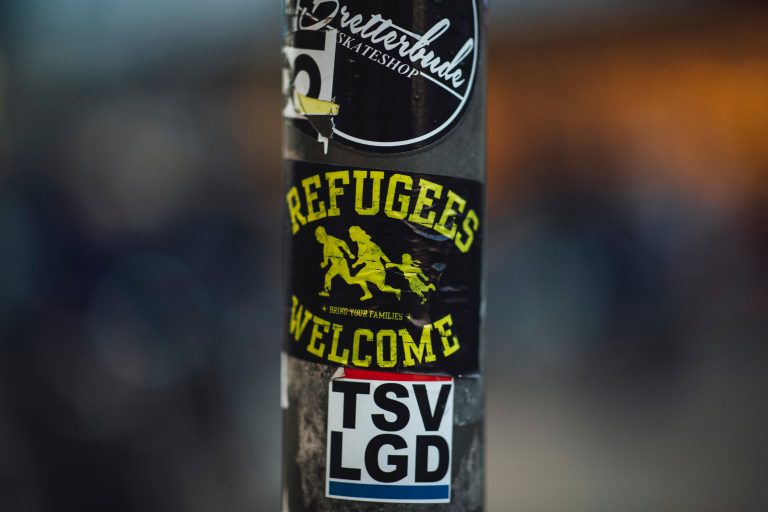 A black and yellow sticker on a telephone pole reads, "Refugees Welcome."