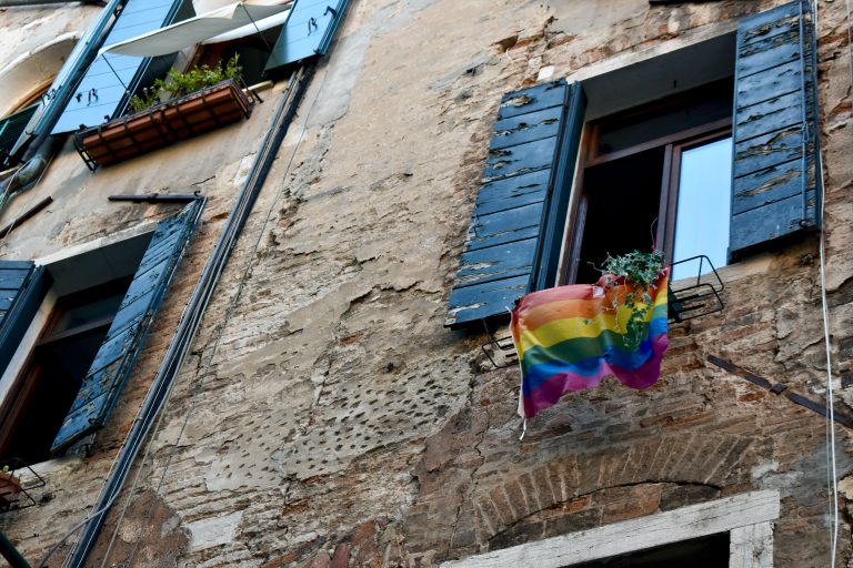A pride flag flies on a window box, on a brown building with green shutters.