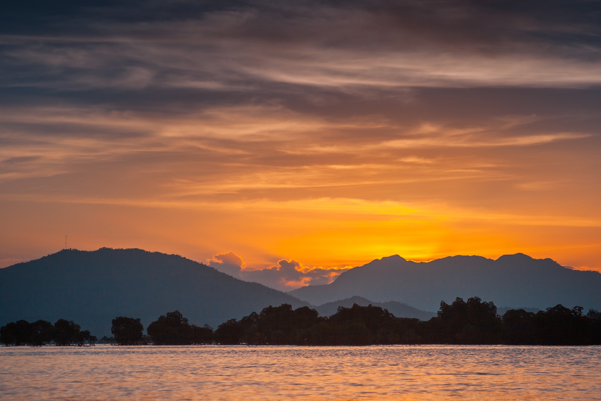 A sun sets serenely behind distant mountains overlooking a lake.