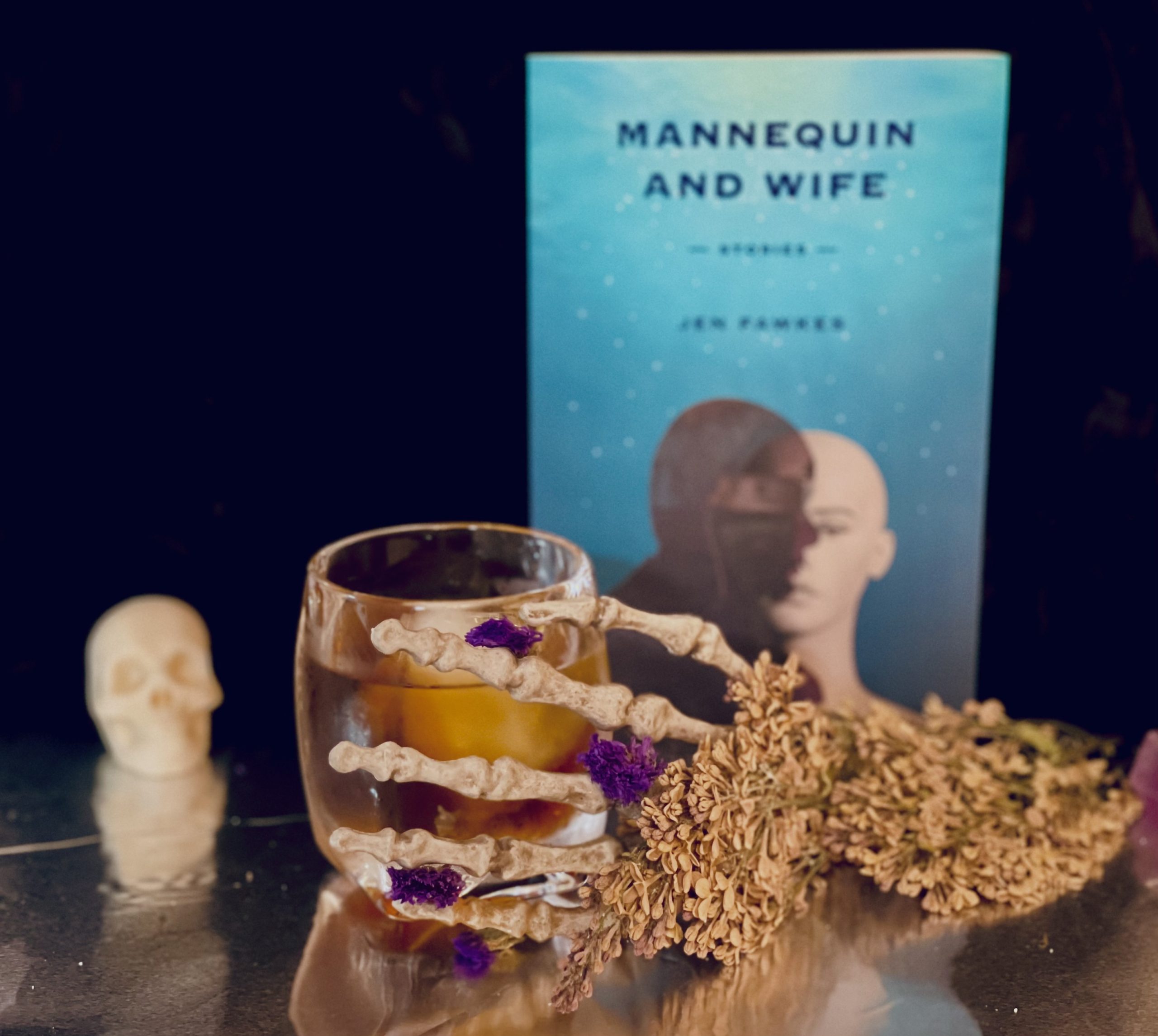 Set against a dark backdrop, a skeleton hand holds a glass containing the cocktail MANNEQUIN AND WIFE, inspired by the book of the same name, which is presented behind it.