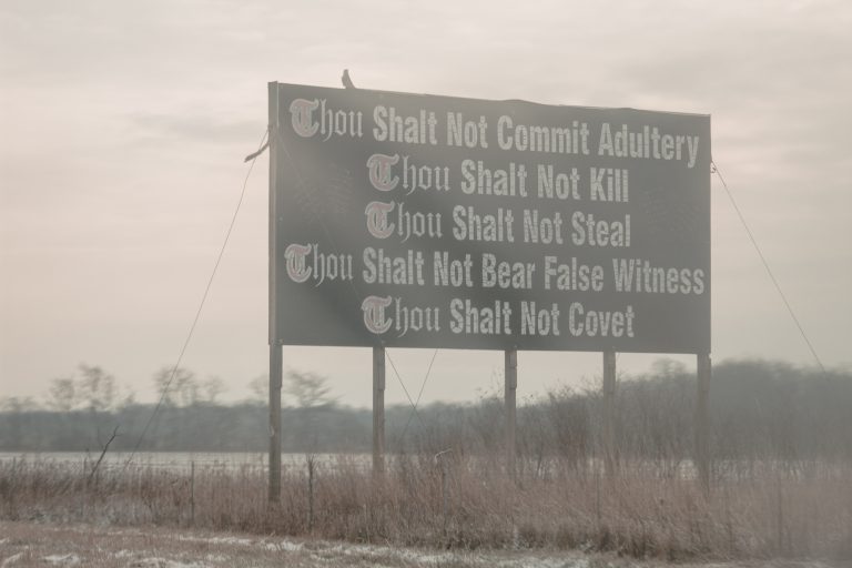 A sign overlooking a foggy country road reads "Thou Shalt Not Commit Adultery"; "Thou Shalt Not Kill"; "Thou Shalt Not Steal"; "Thou Shalt Not Bear False Witness"; "Thou Shalt Not Covet"