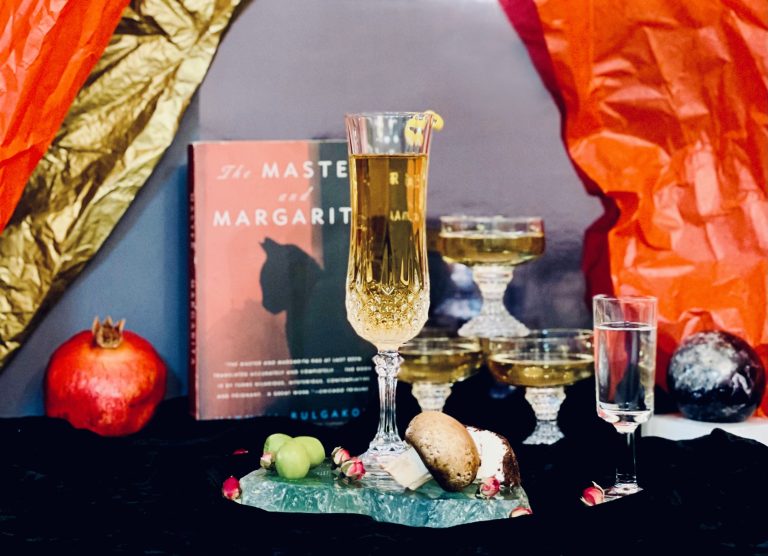 Front and center stands the booktail itself on a slab of fluorite, decorated with olives, a mushroom, soft cheese with a mustard and wine rind, and dried rose buds. Behind it is the book itself, "Master and Margarita". Letters from the book cover behind appear to float in the glass. 