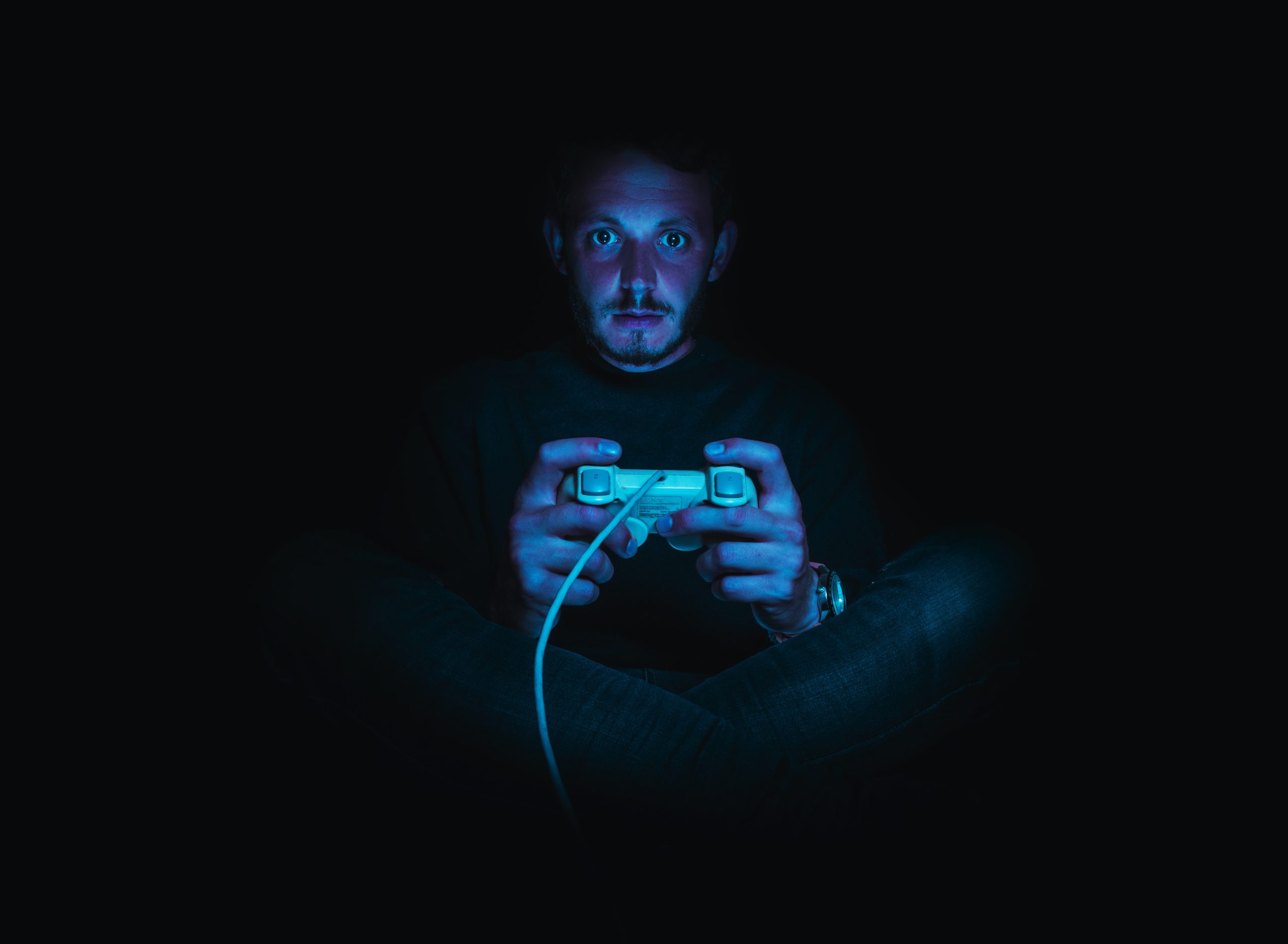 A man sitting in the dark holds a video game controller, staring at the screen.