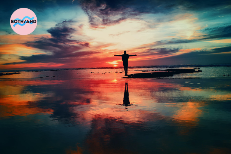 A person stands and watches the sunset with their arms outstretched on a beach.