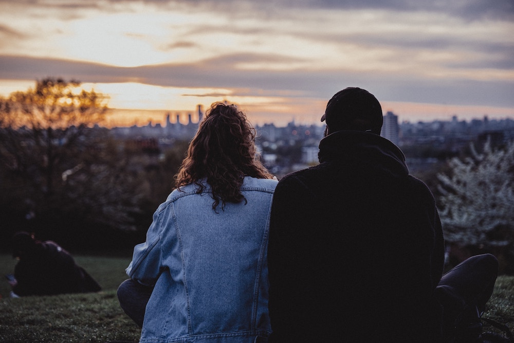 Two people sitting on grass and watching a sunset over the skyline