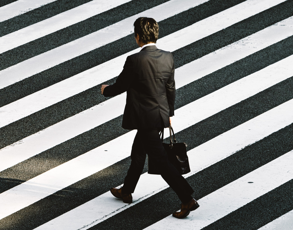 A man in a suit holds a briefcase and walks along a crosswalk.