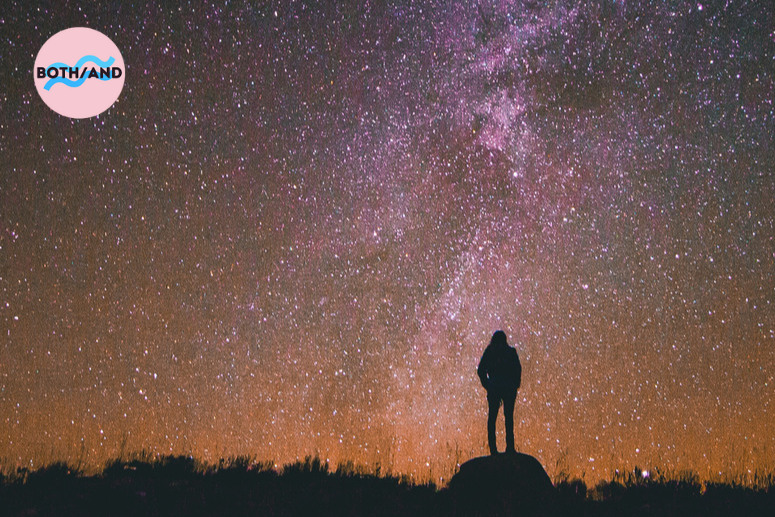 A person stands on top of a rock looking at stars in the night sky.