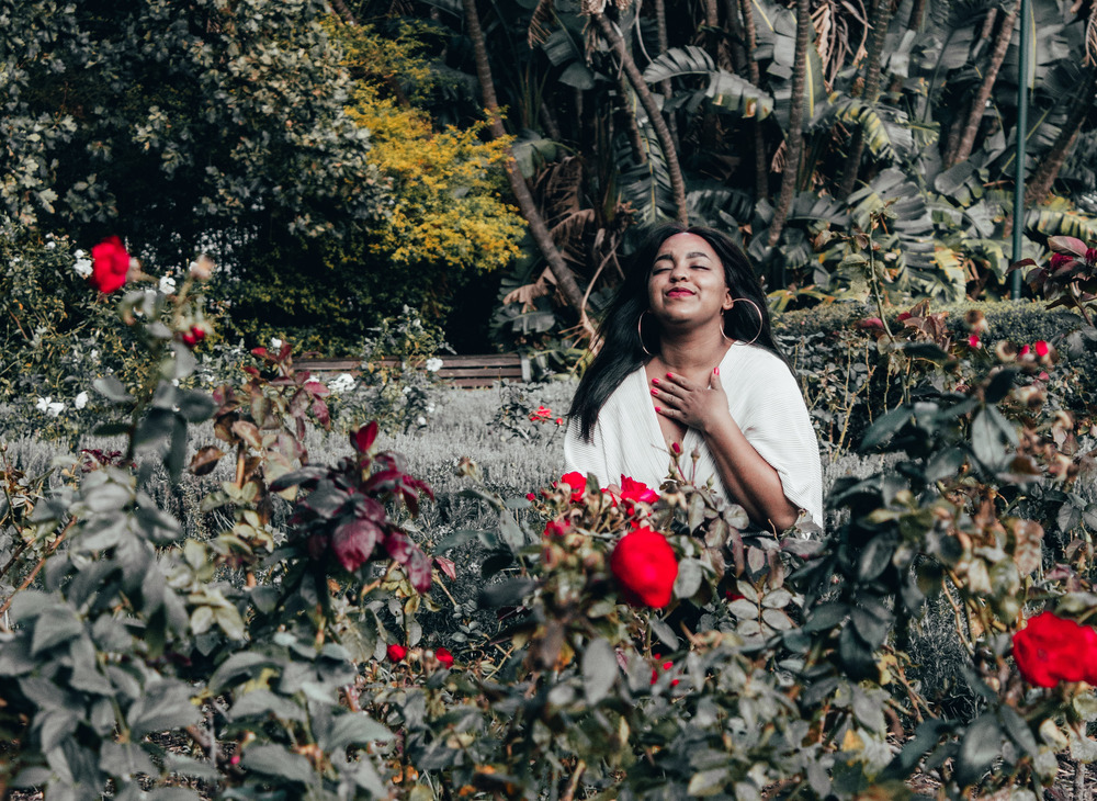 A woman in a white shirt is surrounded by plants and red flowers in a garden in Cape Town, South Africa.