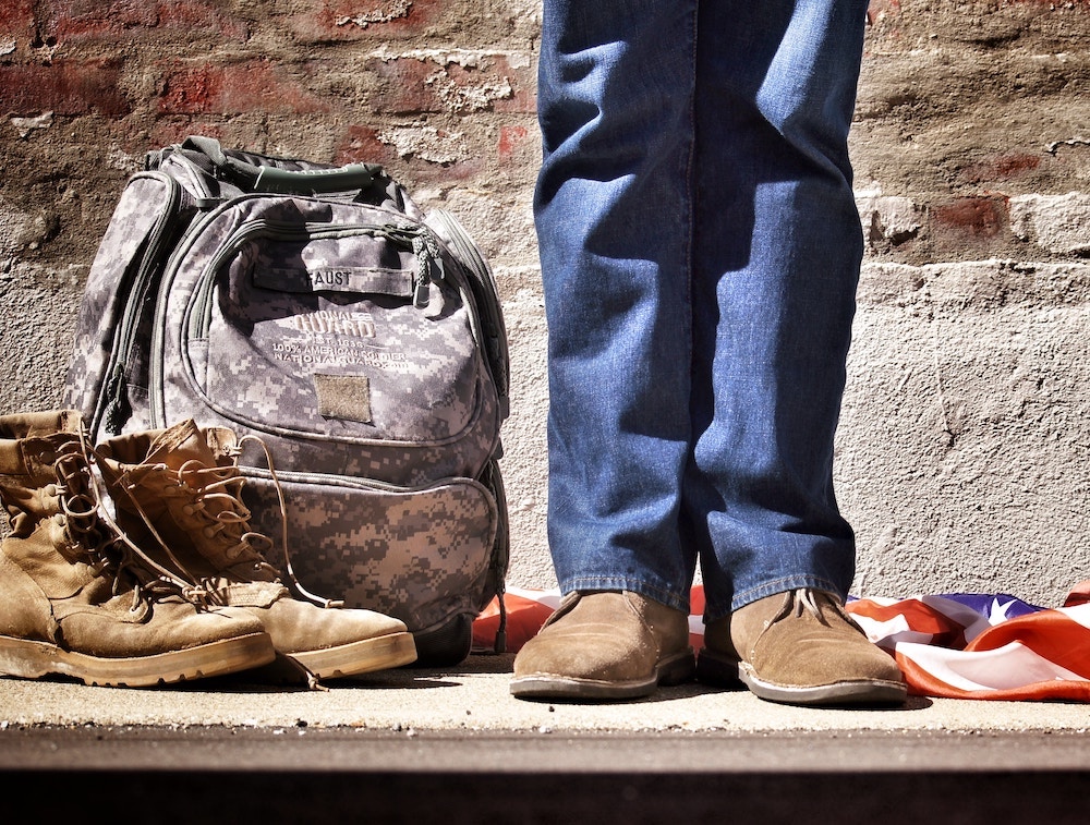 A person stands in civilian boots and shoes next to an army backpack, boots, and an American flag