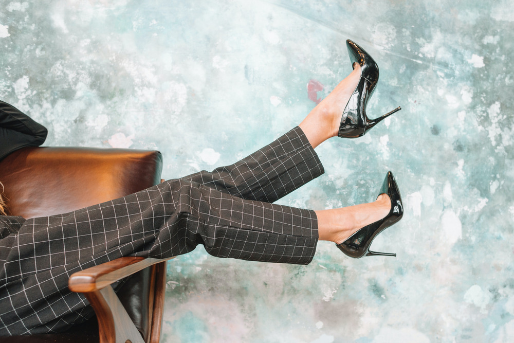 A woman wearing slacks and high heels lounges in a chair, kicking her feet in the air