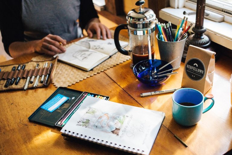 Artist drawing on wooden table full of pens, pencils, coffee
