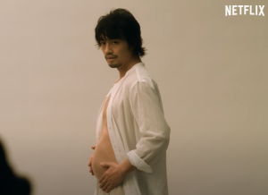 A pregnant man holds his stomach and wears an open white linen shirt.