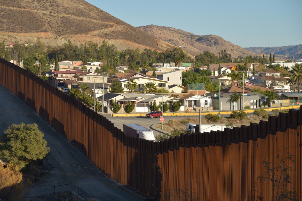 A fence divides the border between America and Tecate, Mexico, with houses visible above the fence.