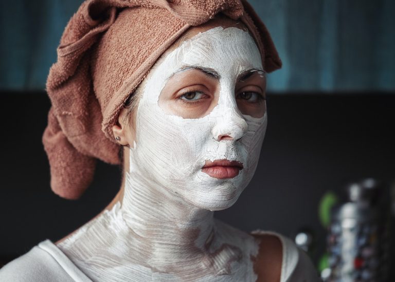 A woman stands with a bath towel wrapped around her head and a face mask all over her face and neck