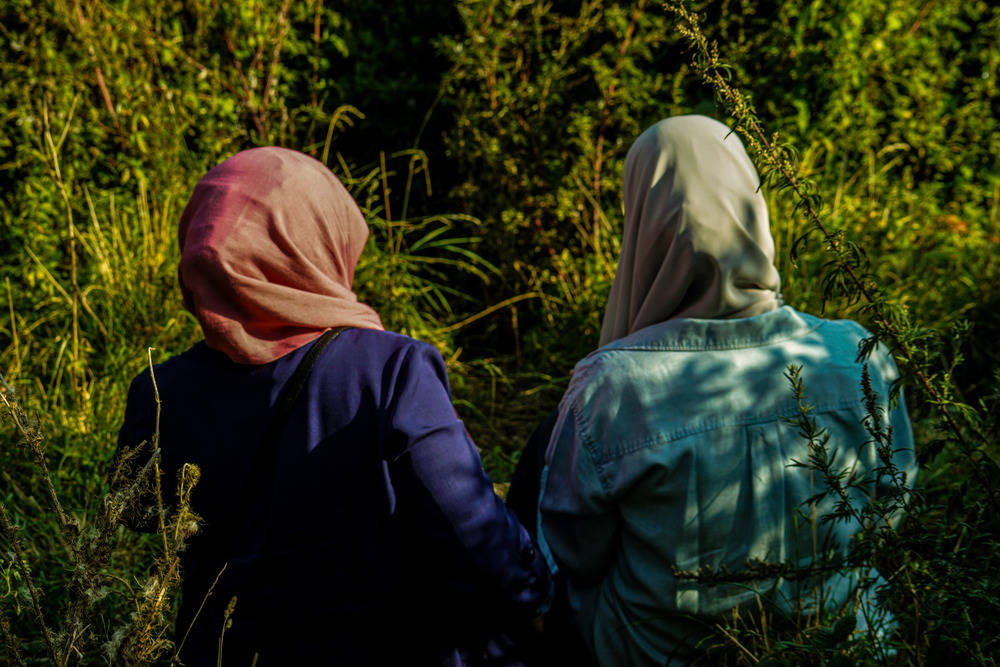 Two women, one wearing a pink hijab and the other a green hijab, sit by a river and are surrounded by greenery