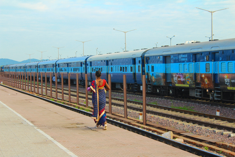A woman in a blue and red sari stands beside a blue train in India