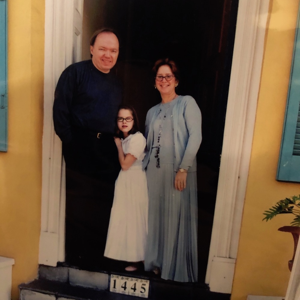 A father, daughter, and mother standing in doorway of yellow house
