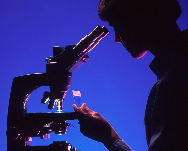 A female pathologist views a slide under a microscope backlit by purple light.