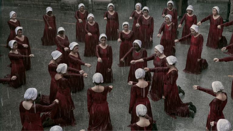 An image from the TV show The Handmaid's Tale where a group of women in red dresses point at one woman in the center of the circle.